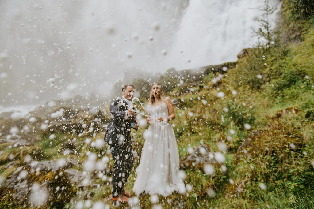 Couple popping champagne under the waterfall after their ceremony during their BC elopement package